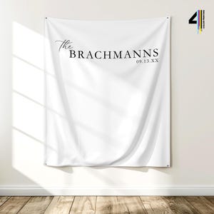 Pu&ograve; includere: Un banner di tessuto bianco con la scritta "The Brachmanns" e la data "09.13.XX" in nero. Il banner &egrave; appeso a una parete bianca sopra un pavimento in legno.