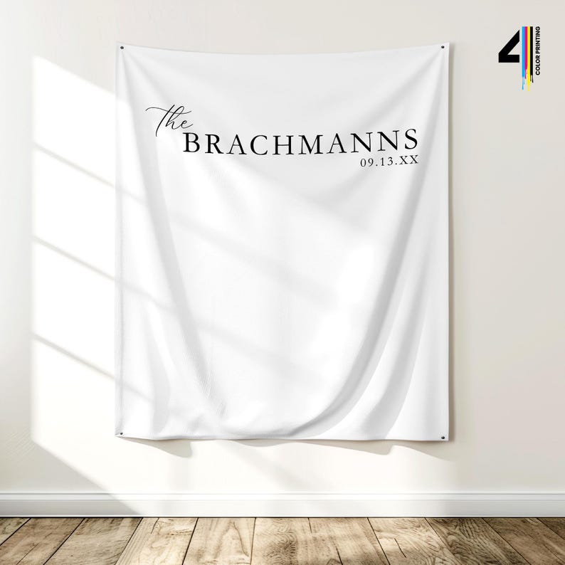 Pu&ograve; includere: Un banner di tessuto bianco con la scritta "The Brachmanns" e la data "09.13.XX" in nero. Il banner &egrave; appeso a una parete bianca sopra un pavimento in legno.