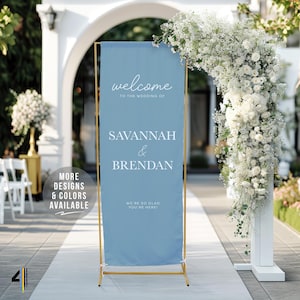 May include: A light blue wedding welcome sign with gold-colored frame. The sign reads "welcome to the wedding of Savannah & Brendan". The sign is displayed outdoors with floral arrangements and an archway.