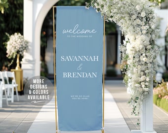 May include: A light blue wedding welcome sign with gold-colored frame. The sign reads "welcome to the wedding of Savannah & Brendan". The sign is displayed outdoors with floral arrangements and an archway.