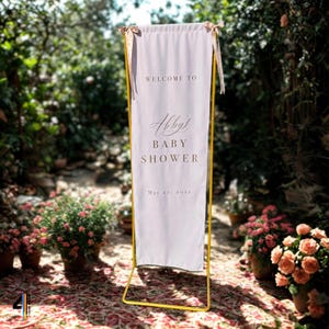 May include: A vertical light pink fabric banner suspended from a gold metal frame. The banner features the text "WELCOME TO", "Abby's", "BABY SHOWER", and "May 22, 20xx" in dark lettering. The top corners are secured with light pink ribbons.