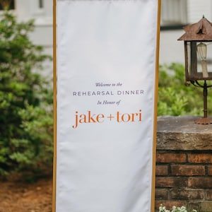 May include: A tall, rectangular white fabric banner held by a light wood frame. The text reads: Welcome to the REHEARSAL DINNER In Honor of jake + tori. The bottom edge features a decorative orange band with a white floral and vine pattern.