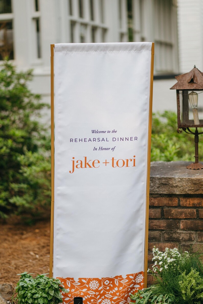 May include: A tall, rectangular white fabric banner held by a light wood frame. The text reads: Welcome to the REHEARSAL DINNER In Honor of jake + tori. The bottom edge features a decorative orange band with a white floral and vine pattern.