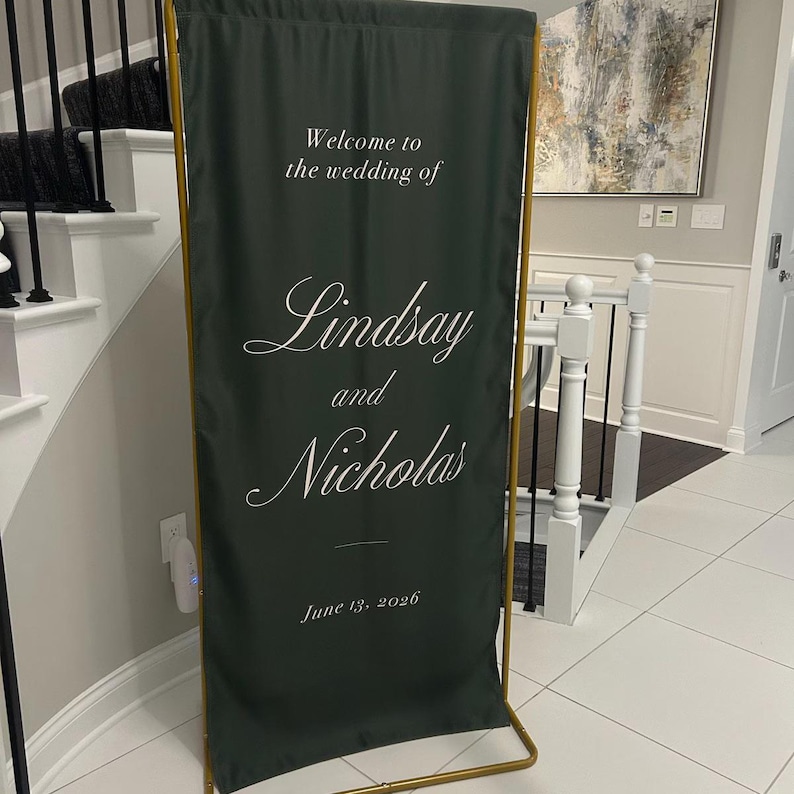 May include: A tall, rectangular dark green fabric wedding welcome sign suspended on a thin gold metal frame. The sign features white printed text that reads: Welcome to the wedding of Lindsay and Nicholas, June 13, 2026, in a mix of serif and script fonts.