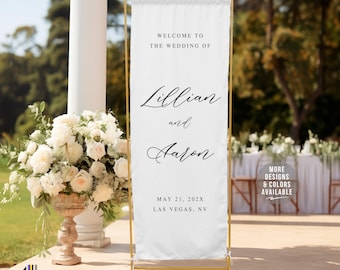 May include: A white wedding welcome sign on a gold stand. The sign reads "Welcome to the Wedding of Lillian and Aaron" with the date and location below. A floral arrangement and outdoor setting are in the background.