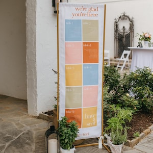 May include: A tall vertical wedding seating chart sign on a gold metal frame. The white fabric banner features the text "we're glad you're here" at the top, a grid of nine colorful rectangular blocks labeled table 1 through 9, and "have a seat" at the bottom.