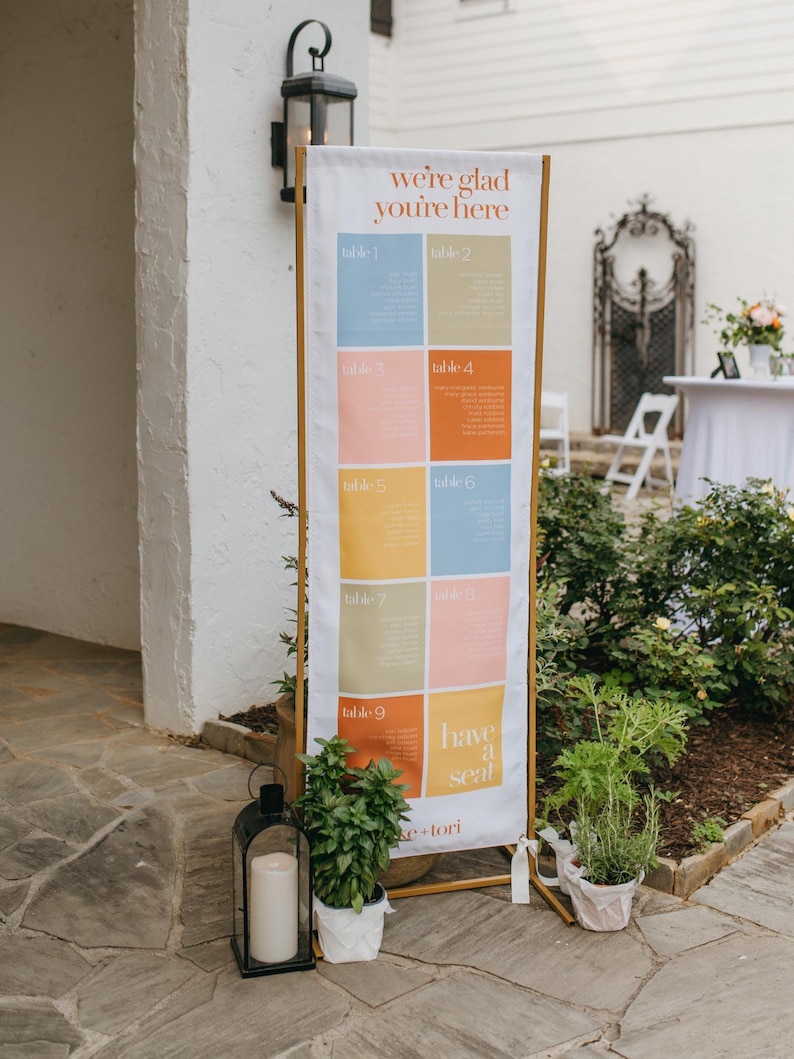 May include: A tall vertical wedding seating chart sign on a gold metal frame. The white fabric banner features the text "we're glad you're here" at the top, a grid of nine colorful rectangular blocks labeled table 1 through 9, and "have a seat" at the bottom.