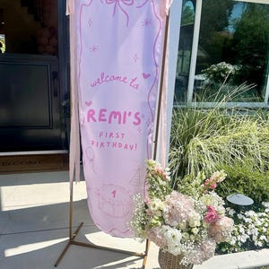 May include: A tall, light pink fabric banner suspended on a thin gold metal frame. The banner features a hand-drawn bow, stars, hearts, and a cake illustration with the text: welcome to REMI'S FIRST BIRTHDAY! Pink ribbons hang from the top corners.