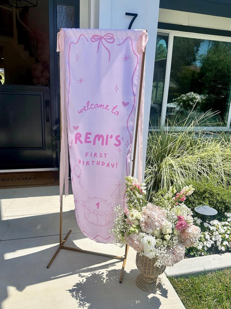 May include: A tall, light pink fabric banner suspended on a thin gold metal frame. The banner features a hand-drawn bow, stars, hearts, and a cake illustration with the text: welcome to REMI'S FIRST BIRTHDAY! Pink ribbons hang from the top corners.