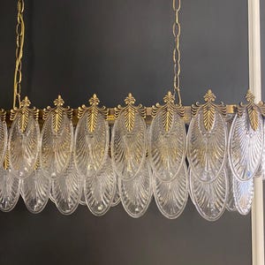 May include: A gold-toned chandelier with clear, oval-shaped glass pendants. The fixture has a rectangular frame with decorative gold accents and is suspended by gold chains. The glass pendants are arranged in a row, creating a sophisticated lighting element.