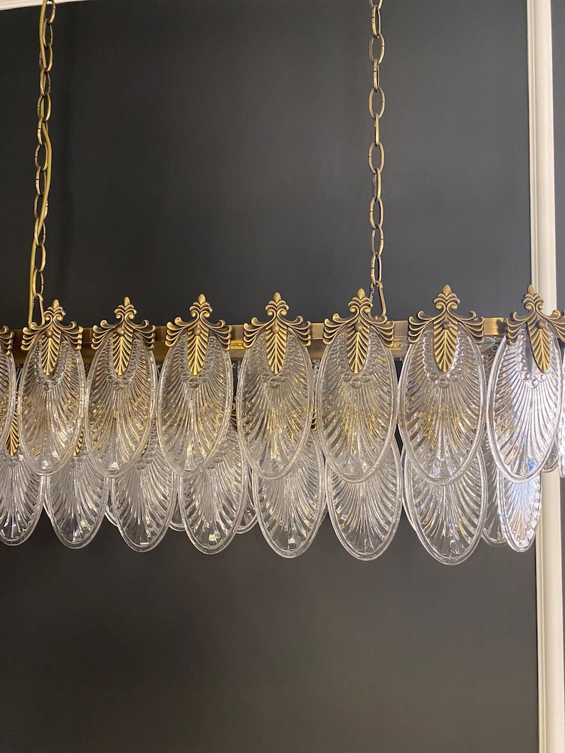 May include: A gold-toned chandelier with clear, oval-shaped glass pendants. The fixture has a rectangular frame with decorative gold accents and is suspended by gold chains. The glass pendants are arranged in a row, creating a sophisticated lighting element.