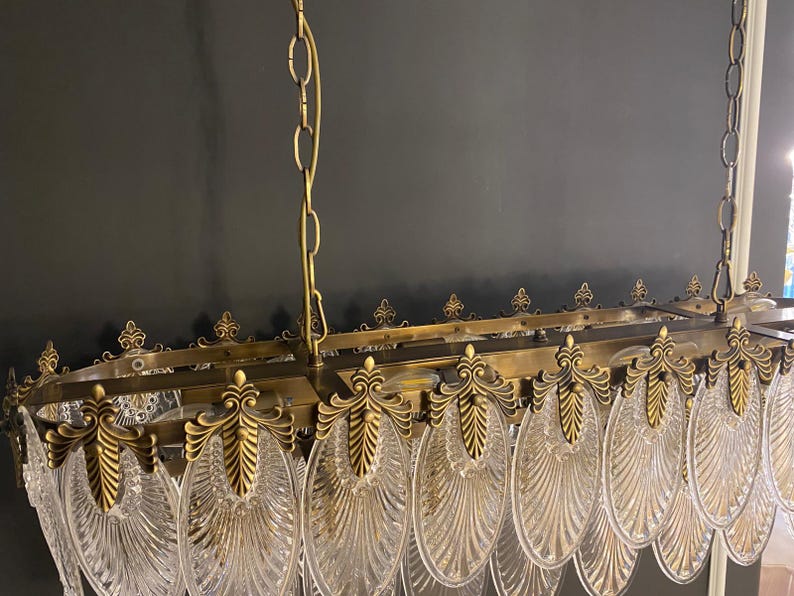 May include: An antique brass chandelier with clear, fan-shaped glass panels. The rectangular frame is adorned with ornate brass detailing and suspended by chains. The design features a vintage aesthetic, suitable for interior lighting.