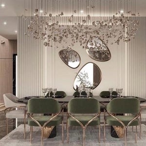 May include: A modern dining room with a large, crystal chandelier, a dark wood table, and six green velvet chairs with gold metal frames. The walls are white with a decorative, organic-shaped mirror.