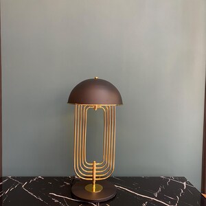 May include: A modern table lamp with a black dome shade and a gold-colored, geometric frame. The lamp sits on a black marble-patterned surface. The lamp's design is a blend of retro and contemporary styles.
