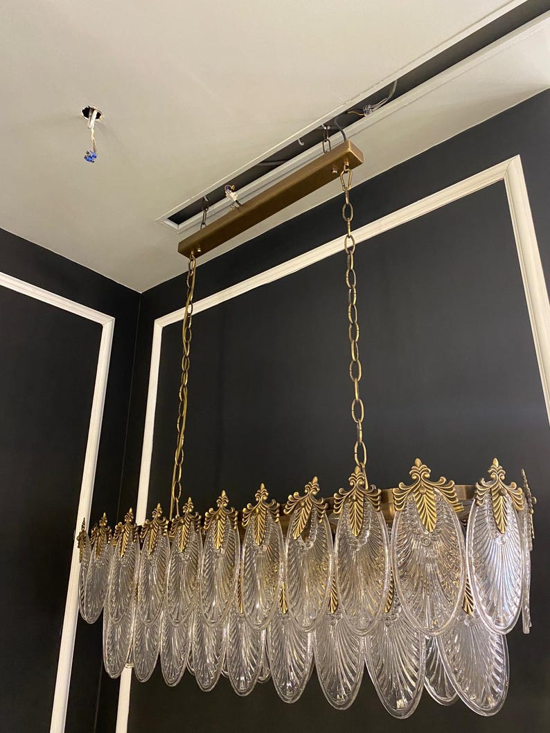 May include: A rectangular chandelier with clear, leaf-shaped glass pendants and gold-colored metal accents. The fixture hangs from a gold-colored metal frame suspended by chains. The design is ornate and elegant, suitable for a dining room or entryway.