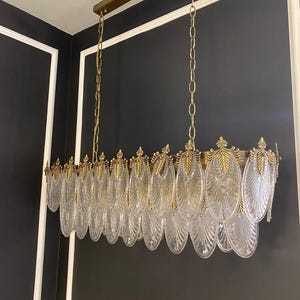 May include: A rectangular chandelier with clear, leaf-shaped glass pendants and gold-colored metal accents. The fixture hangs from a gold-colored metal frame suspended by chains. The design is ornate and elegant, suitable for interior lighting.