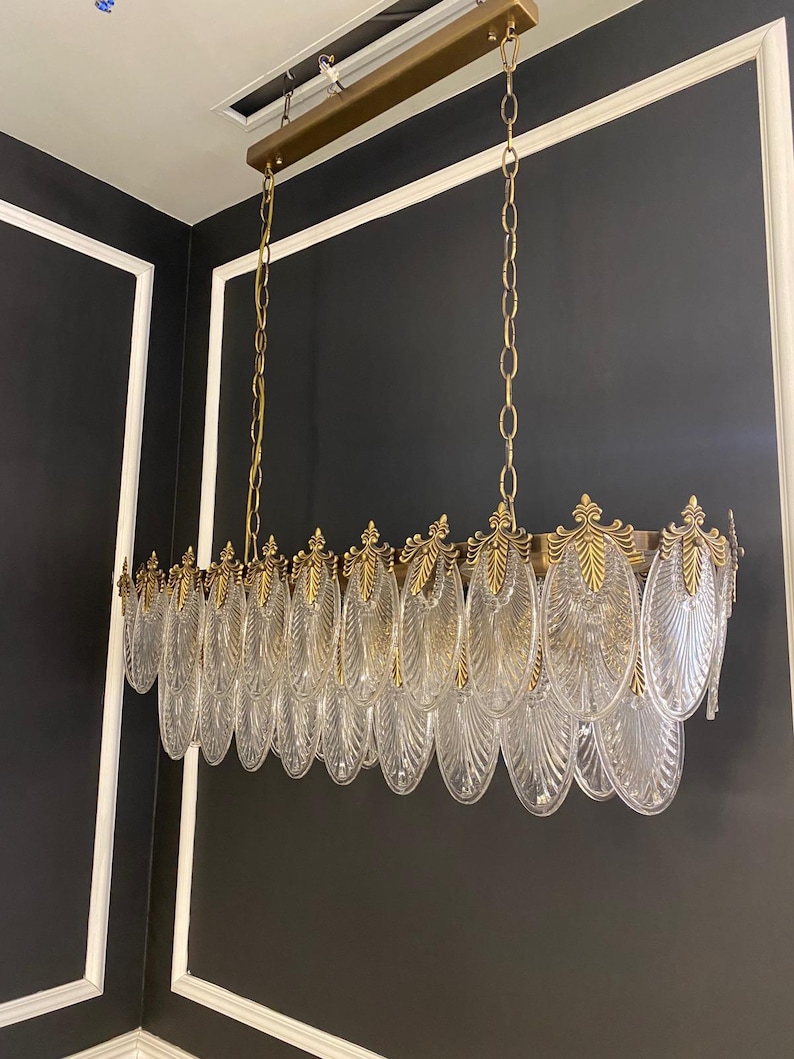 May include: A rectangular chandelier with clear, leaf-shaped glass pendants and gold-colored metal accents. The fixture hangs from a gold-colored metal frame suspended by chains. The design is ornate and elegant, suitable for interior lighting.