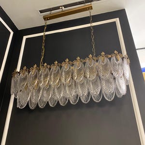 May include: A rectangular chandelier with a gold-colored frame and chain. The chandelier features multiple clear, oval-shaped glass elements, creating a layered, decorative design. The fixture is suspended from the ceiling against a black wall with white trim.