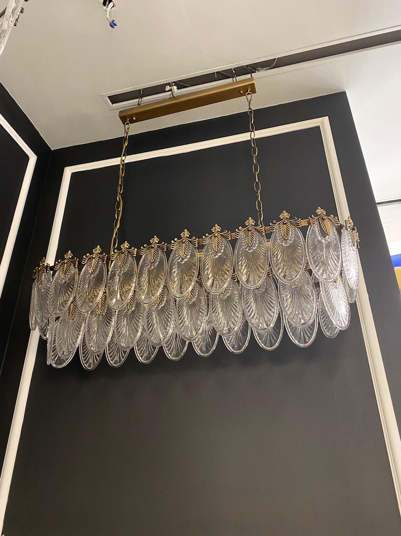May include: A rectangular chandelier with a gold-colored frame and chain. The chandelier features multiple clear, oval-shaped glass elements, creating a layered, decorative design. The fixture is suspended from the ceiling against a black wall with white trim.