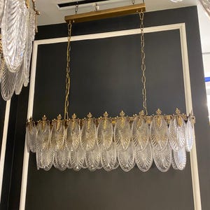 May include: A rectangular chandelier with clear, leaf-shaped glass pendants and gold-colored metal accents. The fixture hangs from gold chains and a gold-colored metal bar. The background is a dark gray wall with white trim.
