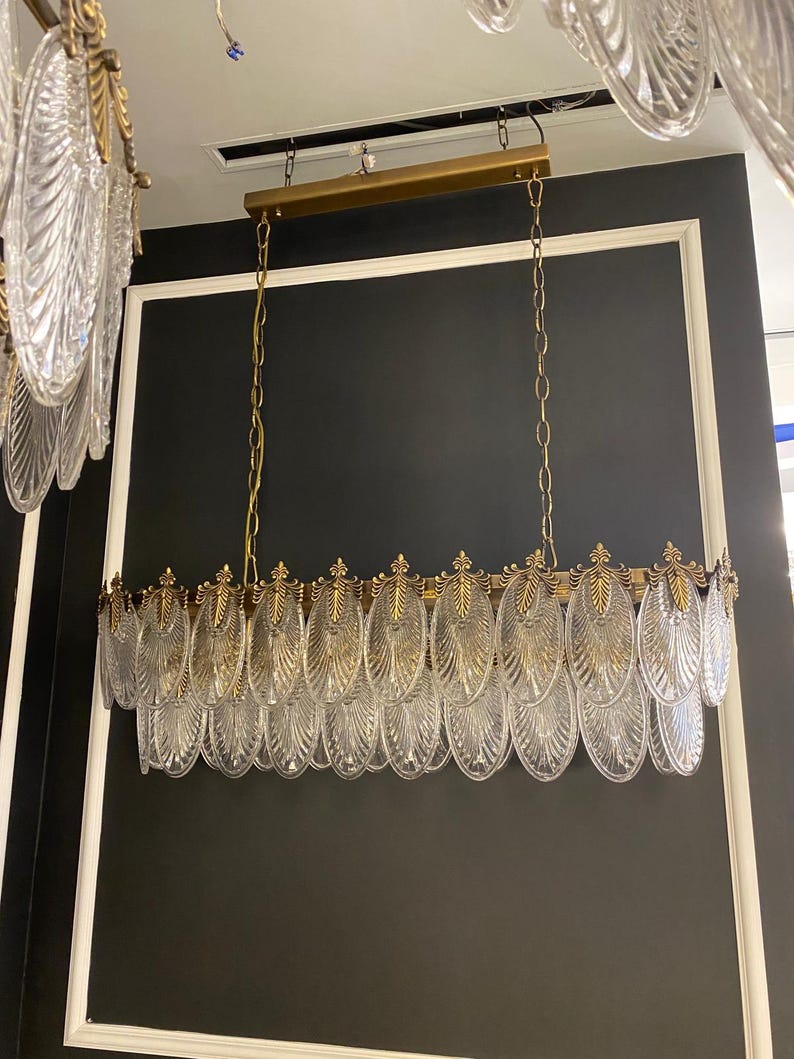 May include: A rectangular chandelier with clear, leaf-shaped glass pendants and gold-colored metal accents. The fixture hangs from gold chains and a gold-colored metal bar. The background is a dark gray wall with white trim.