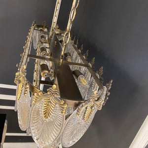 May include: An ornate chandelier with a brass frame and clear, leaf-shaped glass panels. The fixture is suspended from a chain and features decorative gold accents. The chandelier is mounted on a dark gray ceiling.