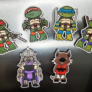 Op de afbeelding: Set van zes magneten met Teenage Mutant Ninja Turtles en Shredder-personages. De magneten zijn gemaakt van hout en hebben een cartoon-stijl. De personages zijn geschilderd in hun kenmerkende kleuren, waaronder groen, rood, blauw, paars en bruin.