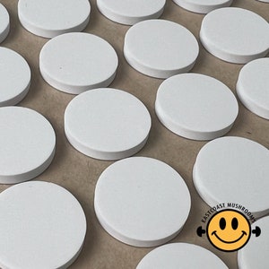 May include: A collection of white round discs, possibly coasters or placemats, arranged in rows on a brown surface. The discs are smooth and have a slightly glossy finish.