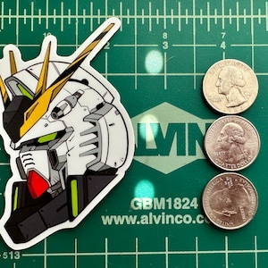 May include: A white, green, and red anime robot sticker with a yellow and black design. The sticker is on a green cutting mat with a ruler and three US quarters.