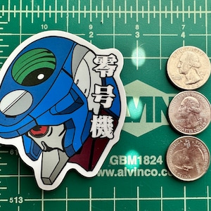 May include: A blue and red sticker with a cartoon robot head and Japanese text that reads "Zero Unit". The sticker is on a green cutting mat with a ruler and US currency.
