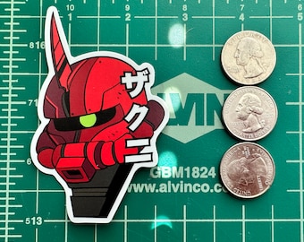 May include: A red and black sticker featuring a stylized robot head with Japanese text. The sticker is next to three quarters on a green cutting mat. The robot head has a prominent antenna and a green eye.