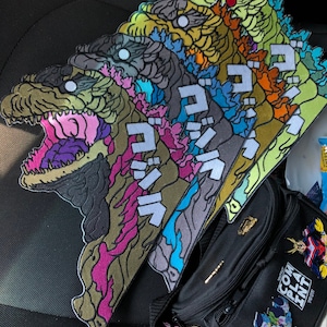 May include: A set of seven colorful embroidered patches featuring a Godzilla-like monster with Japanese text. The patches are arranged in a row, with the largest patch at the bottom and the smallest patch at the top. The patches are all different colors, including green, blue, purple, pink, and yellow.
