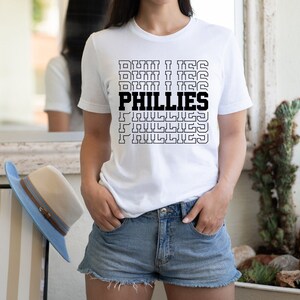 May include: White t-shirt with a black graphic print of the word "PHILLIES" repeated in a stacked pattern. The graphic is in a retro style with a black outline.