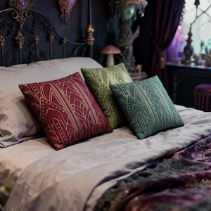 Może przedstawiać: Trzy kwadratowe poduszki dekoracyjne w kolorze bordowym, zielonym i turkusowym ze złotymi geometrycznymi wzorami leżą na łóżku. Poduszki dodają elegancji wystrojowi sypialni. Łóżko ma szarą kołdrę i ciemne wezgłowie.