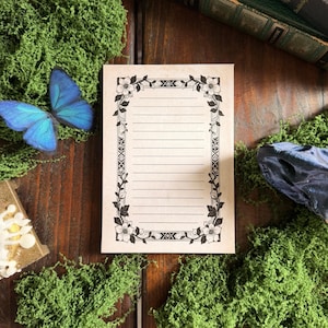 May include: A rectangular piece of stationery paper featuring a black ink floral border design with horizontal lines for writing. The paper has a light beige tone and is surrounded by green moss, a blue butterfly, and a dark book.