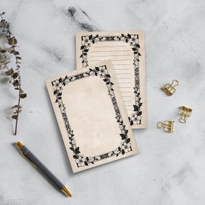 Op de afbeelding: Twee beige notitieblokken met zwarte bloemen en geometrische randen. Eén notitieblok heeft gelinieerde pagina's, de andere is blanco. Een grijze pen met een gouden punt en drie gouden paperclips zijn ook zichtbaar.