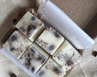 May include: Six white wax melts with a brown coffee bean swirl pattern are arranged in a cardboard box. The box is open and the wax melts are visible. The box is sitting on a beige fabric surface.