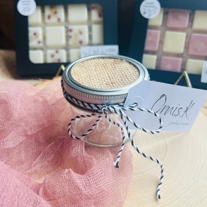 Op de afbeelding: Een klein glazen potje met een deksel van jute en een wit label met de tekst "OrvisK Candle Co.". Het potje is vastgebonden met een zwart-wit touwtje en staat op een houten oppervlak met een roze stof op de achtergrond.