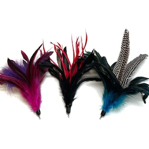 May include: Three feather cat toys with different colored feathers. The first toy has purple, pink, and black feathers. The second toy has red, black, and white feathers. The third toy has blue, black, and white feathers.
