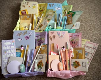 May include: Four gift boxes filled with stationery items including notebooks, pens, pencils, lip balms, tea bags, and decorative cards. Each box features a color-coordinated theme with items in shades of yellow, blue, purple, and pink, featuring various floral, fruit, and religious motifs.