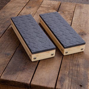 May include: Two rectangular wooden foot rests with a black, textured top. The foot rests are made of light-colored wood and are secured with small metal screws. They are placed on a rustic wooden table.