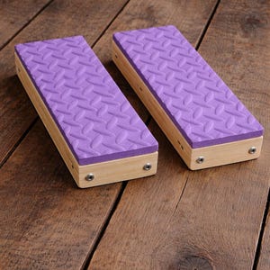 May include: Two rectangular wooden blocks with a light purple, textured top. The blocks are made of light-colored wood and are secured with small silver screws. The blocks are placed on a wooden surface.