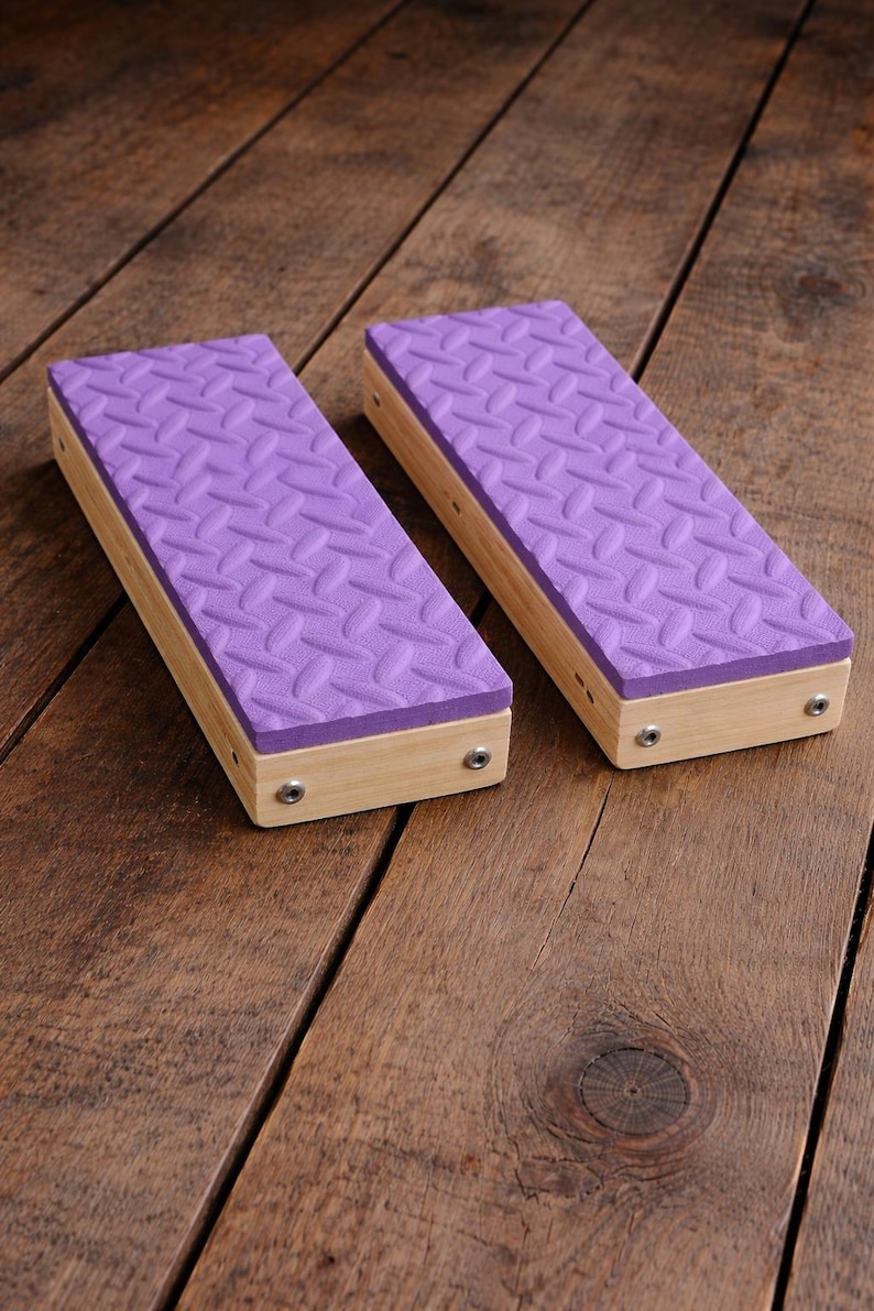 May include: Two rectangular wooden blocks with a light purple, textured top. The blocks are made of light-colored wood and are secured with small silver screws. The blocks are placed on a wooden surface.