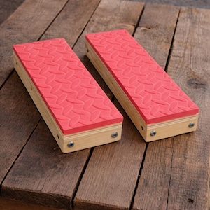 May include: Two wooden foot blocks with red, textured tops. The blocks are rectangular, with light-colored wood sides and a pattern of ovals on the red surface. Metal screws secure the top to the wooden base. The blocks are on a rustic wooden surface.