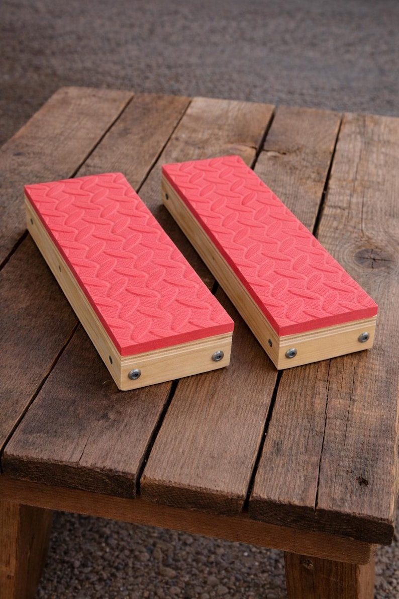 May include: Two wooden foot blocks with red, textured tops. The blocks are rectangular, with light-colored wood sides and a pattern of ovals on the red surface. Metal screws secure the top to the wooden base. The blocks are on a rustic wooden surface.