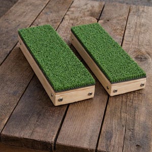 May include: Two rectangular wooden blocks with a vibrant green, textured surface, resembling artificial turf. The blocks are made of light-colored wood and are secured with small metal fasteners. They are displayed on a rustic wooden table.