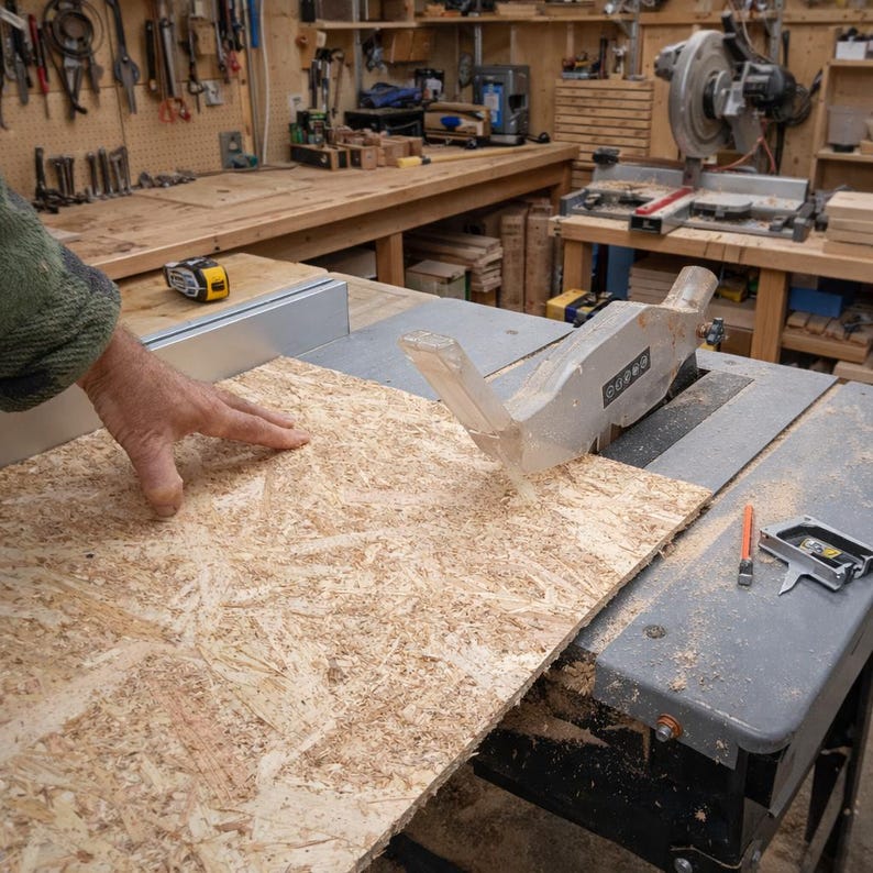 May include: A table saw cutting a sheet of oriented strand board (OSB). The OSB is a light brown color with visible wood chips. A hand is guiding the wood through the saw blade. The saw is gray and silver.