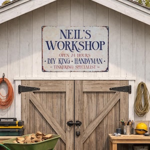 May include: A weathered metal sign reading "NEIL'S WORKSHOP" in navy blue lettering, mounted on a white wooden shed. The sign also states "OPEN 24 HOURS" and lists "DIY KING - HANDYMAN - TINKERING SPECIALIST". The shed doors are made of wood.