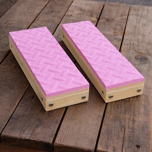 May include: Two rectangular wooden blocks with pink, textured tops. The pink surface has a repeating pattern. The blocks are made of light-colored wood and are secured with small metal screws. The blocks are on a weathered wooden surface.
