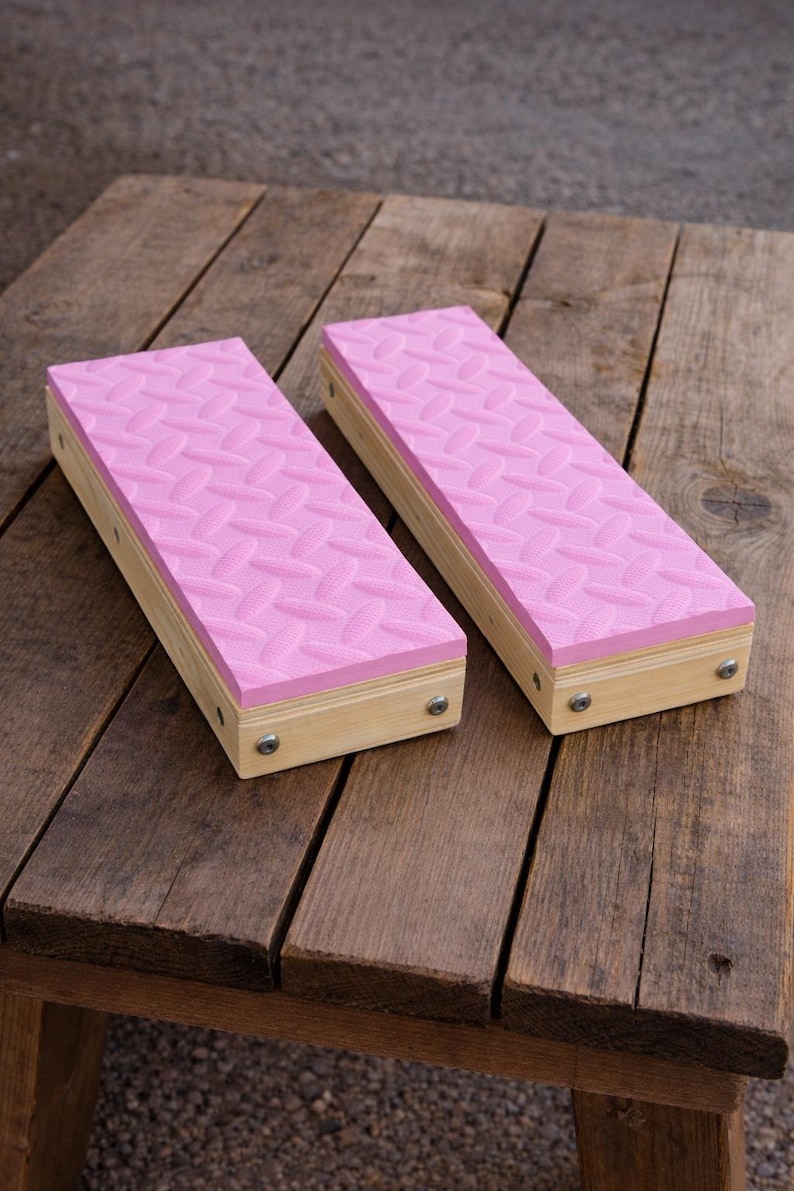 May include: Two rectangular wooden blocks with pink, textured tops. The pink surface has a repeating pattern. The blocks are made of light-colored wood and are secured with small metal screws. The blocks are on a weathered wooden surface.